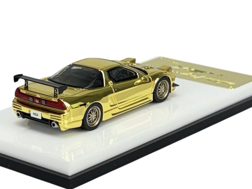 Złoty model samochodu w skali 1:64 z widocznym napisem \'NSX\' na tablicy rejestracyjnej, umieszczony na białej podstawce.