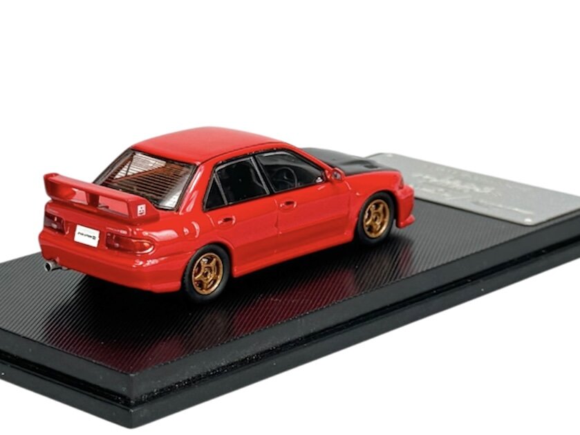 Czerwony model samochodu w skali 1:64 umieszczony na czarnej podstawce. Samochód ma złote felgi i widoczny tylny spojler.