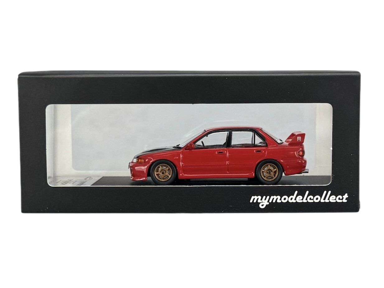 Model czerwonego samochodu w skali 1:64 znajduje się w czarnym pudełku z przezroczystym frontem. Na pudełku widnieje napis \'mymodelcollect\'.
