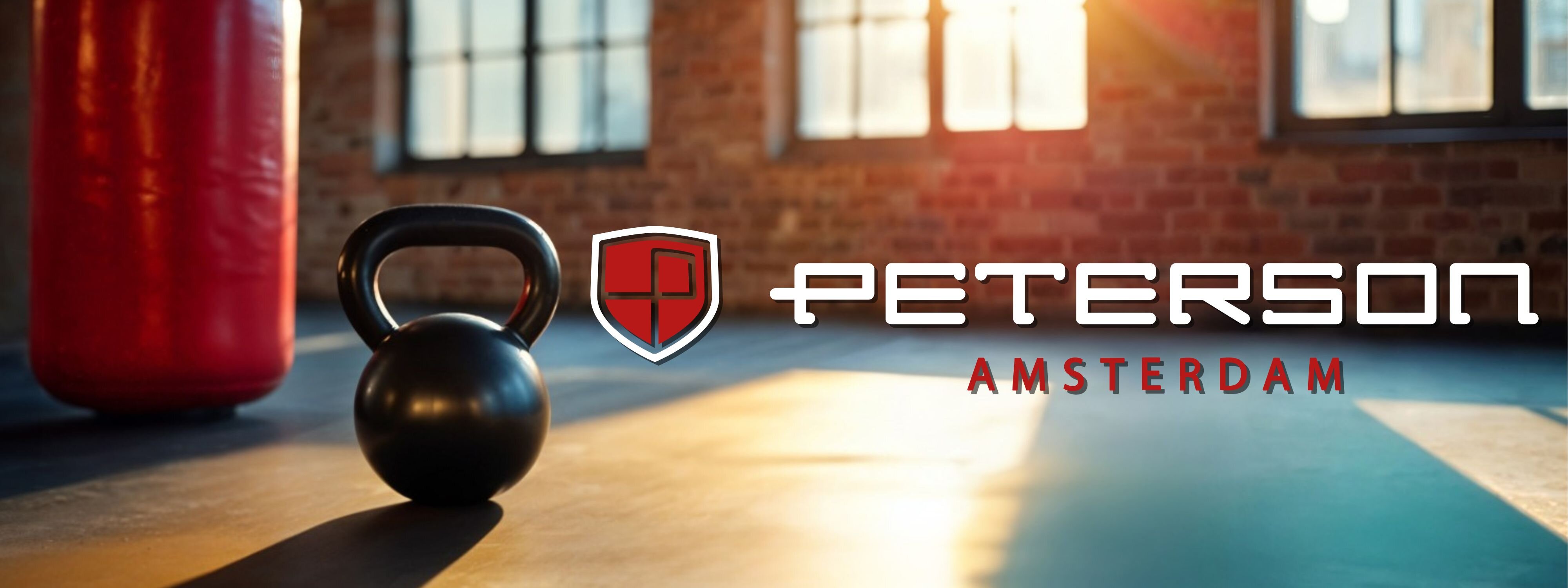Na podłodze w jasnym pomieszczeniu z ceglaną ścianą i dużymi oknami znajduje się czarny kettlebell. Obok widoczne jest logo w kształcie tarczy oraz napis \'PETERSON AMSTERDAM\'.
