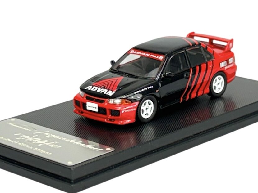 Model samochodu w skali 1:64 przedstawia czerwono-czarny pojazd z napisem \'ADVAN\' na masce. Samochód umieszczony jest na czarnej podstawce z metalową tabliczką.