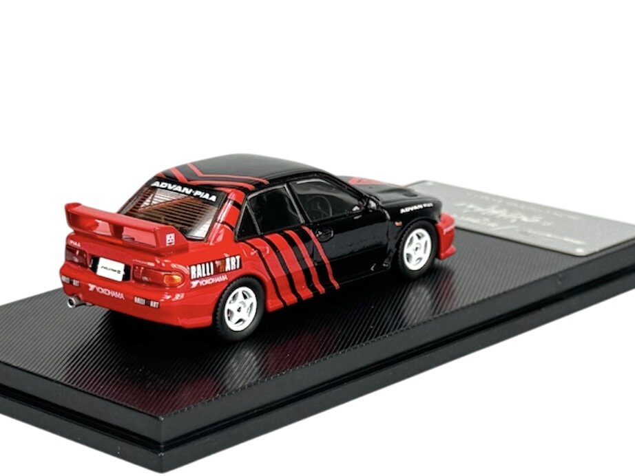 Model samochodu w skali 1:64 w kolorze czerwono-czarnym z napisami \'ADVAN\' i \'RALLI ART\' na tylnej części. Samochód umieszczony jest na czarnej podstawce.