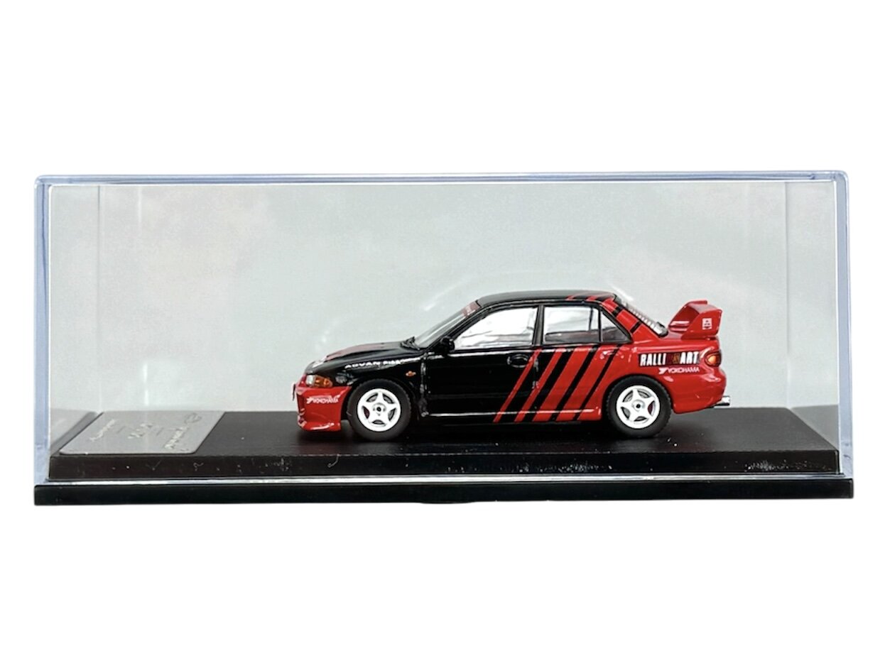 Model samochodu w skali 1:64 w kolorze czarno-czerwonym z widocznymi napisami \'RALLI ART\' i \'YOKOHAMA\' na boku. Samochód umieszczony jest w przezroczystej gablocie na czarnej podstawce.