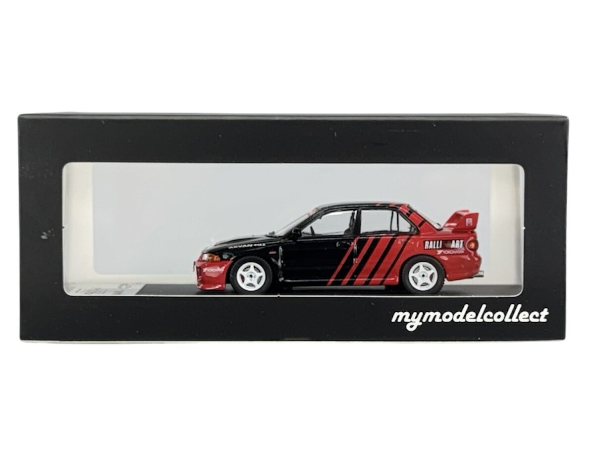 Model samochodu w skali 1:64 w kolorze czarno-czerwonym z białymi felgami, umieszczony w czarnym pudełku z przezroczystym frontem. Na pudełku widnieje napis \'mymodelcollect\'.