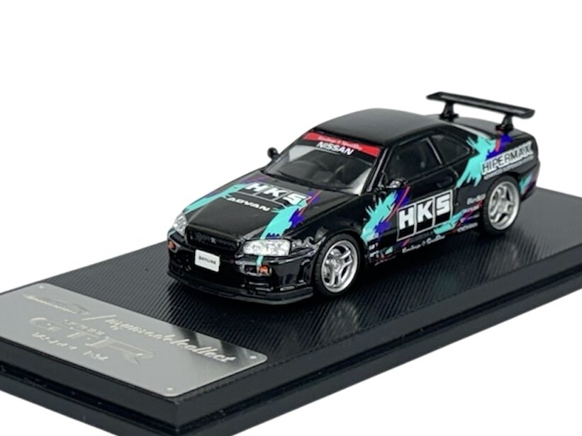 Model samochodu w skali 1:64 przedstawia czarny pojazd z kolorowymi grafikami i napisami \'HKS\' oraz \'NISSAN\' na przedniej szybie. Samochód umieszczony jest na podstawce z napisem \'Nissan Skyline GT-R\'.