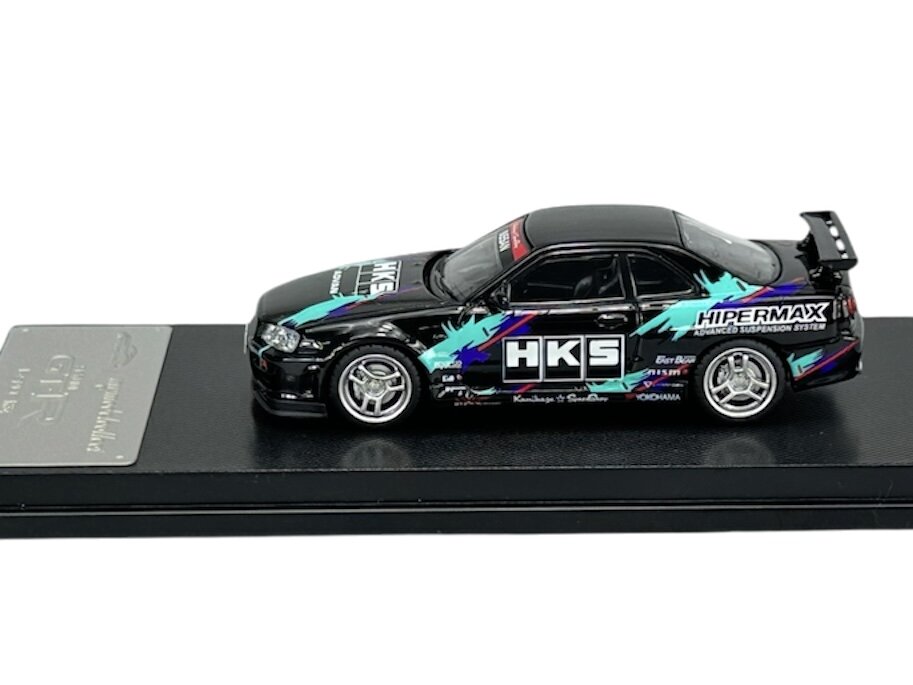 Model samochodu w skali 1:64 przedstawia czarny pojazd z kolorowymi grafikami i napisami \'HKS\' oraz \'HIPERMAX\'. Samochód umieszczony jest na czarnej podstawce z metalową tabliczką.