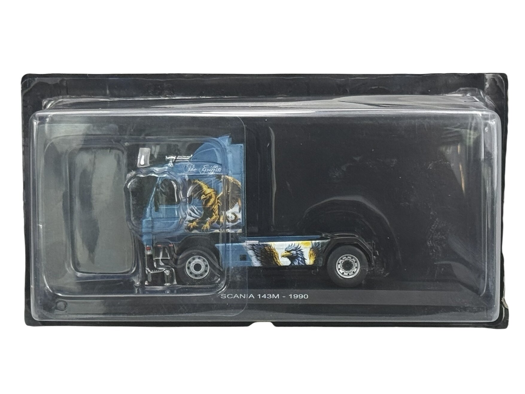 Model ciężarówki w skali 1:43 w opakowaniu, z widocznym napisem \'Scania 143M - 1990\'. Kabina jest niebieska z grafiką przedstawiającą gryfa.