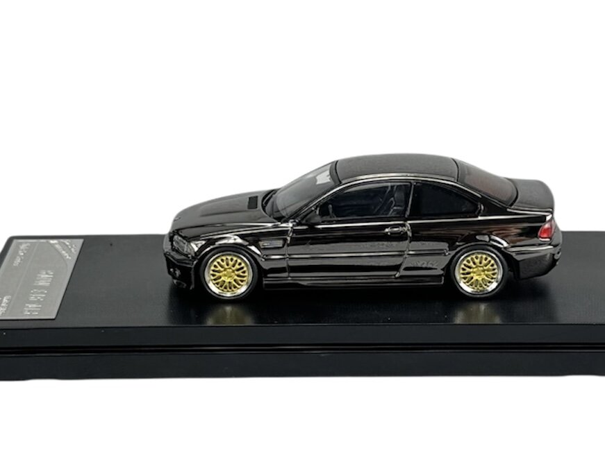 Model samochodu w kolorze ciemnym z złotymi felgami, umieszczony na czarnej podstawce. Na podstawce widoczny jest napis \'BMW M3 E46\'.
