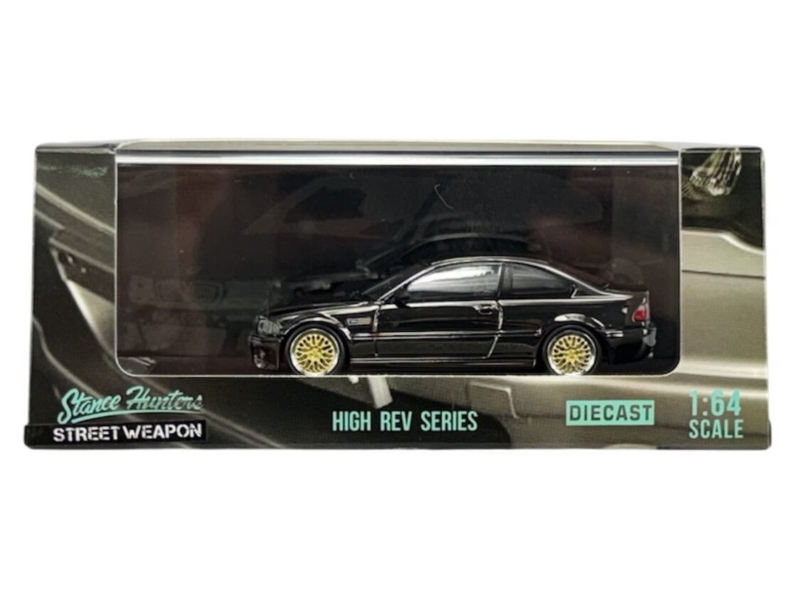 Model samochodu w skali 1:64 w kolorze czarnym z żółtymi felgami, umieszczony w przezroczystej gablotce. Na opakowaniu widnieje napis: \'Stance Hunters STREET WEAPON HIGH REV SERIES DIECAST 1:64 SCALE\'.