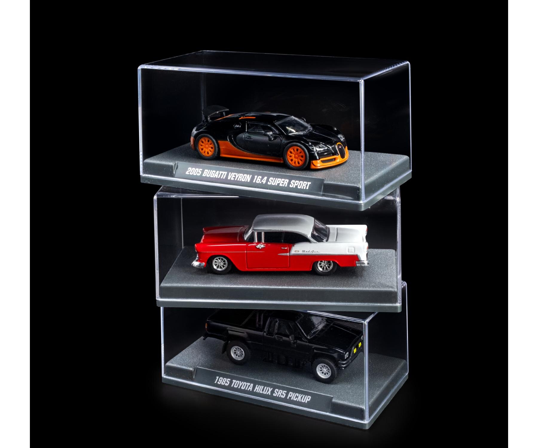Na obrazie widoczne są trzy modele samochodów w przezroczystych gablotach. Pod każdym modelem znajduje się napis: \'2005 Bugatti Veyron 16.4 Super Sport\', \'1955 Chevrolet Bel Air\', \'1985 Toyota Hilux SR5 Pickup\'.