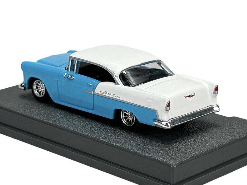 Model samochodu w skali 1:64 przedstawia niebiesko-biały Chevrolet Bel Air z 1955 roku, umieszczony na czarnej podstawce. Na boku pojazdu widoczny jest napis \'Bel Air\'.