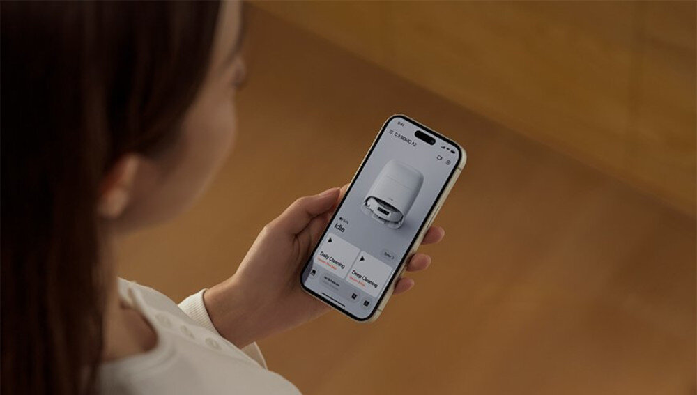Osoba trzyma smartfon, na którym wyświetlana jest aplikacja z obrazem robota sprzątającego. Na ekranie widoczne są przyciski i teksty, ale nie są one czytelne.