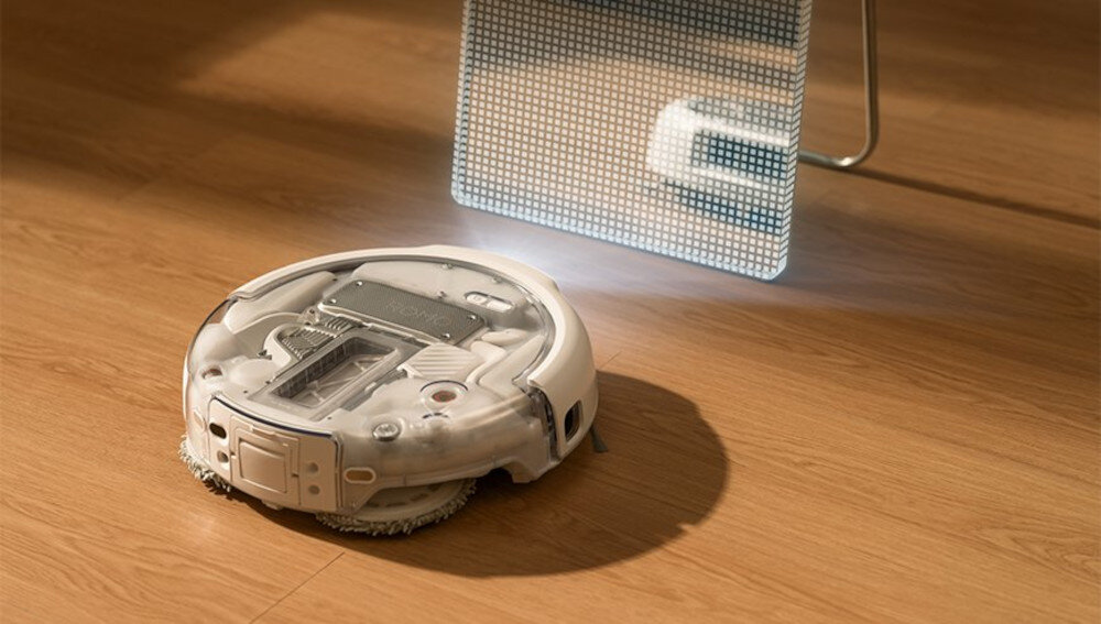 Na drewnianej podłodze znajduje się biały robot sprzątający o okrągłym kształcie. W tle widoczna jest jasna, perforowana powierzchnia z delikatnym podświetleniem.