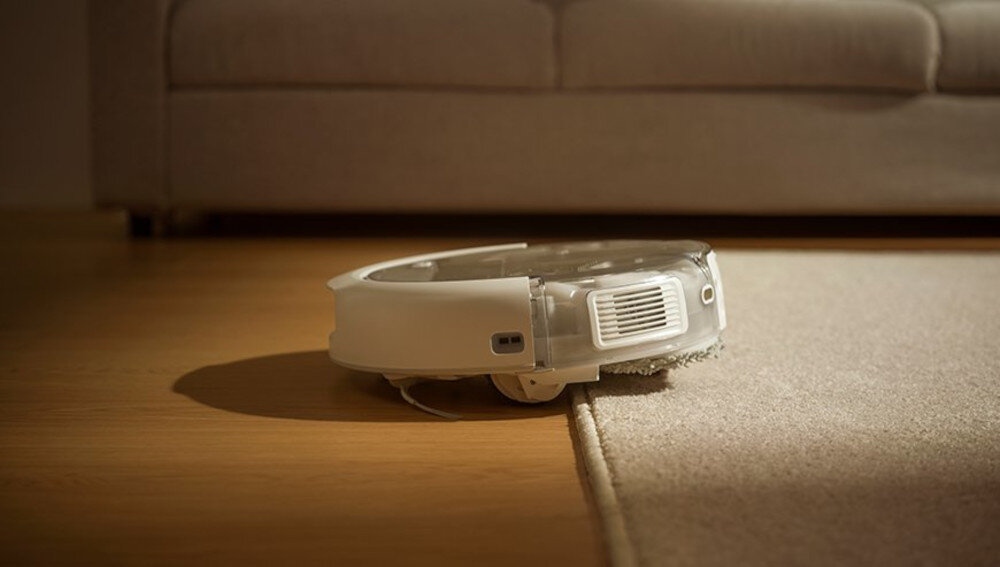 Robot sprzątający na podłodze, częściowo na dywanie, w tle widoczna kanapa.