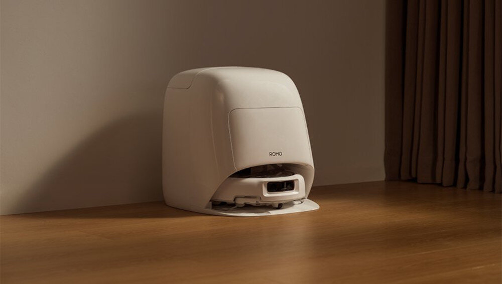 Na drewnianej podłodze stoi biały robot sprzątający z widocznym napisem \'ROMO\'. W tle znajduje się zasłona.