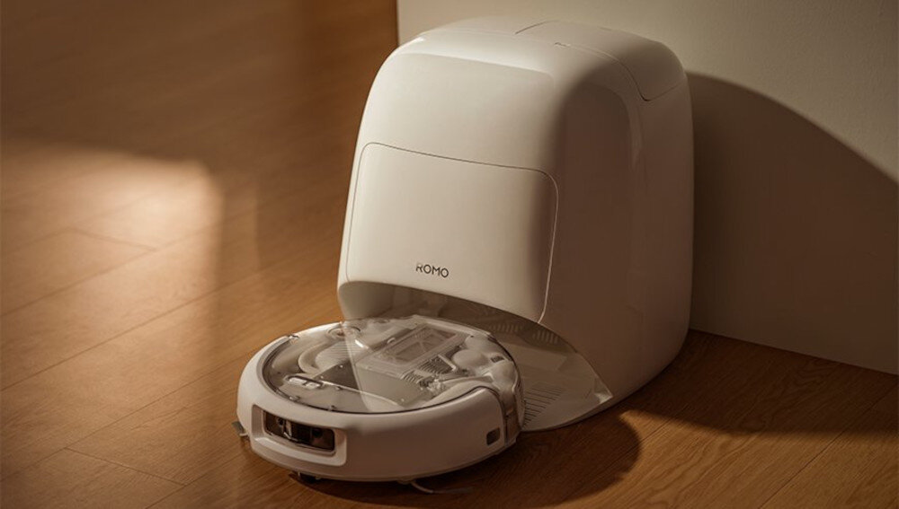 Na drewnianej podłodze znajduje się biały robot sprzątający z napisem \'ROMO\' na stacji dokującej.