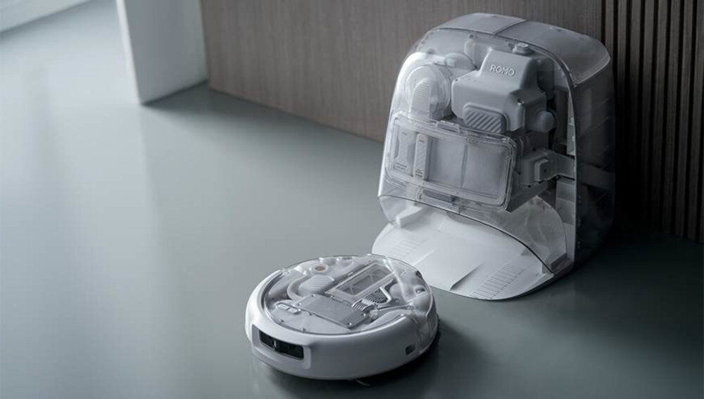 Na obrazie widoczny jest biały robot sprzątający oraz jego stacja dokująca. Na stacji znajduje się napis \'ROMO\'.