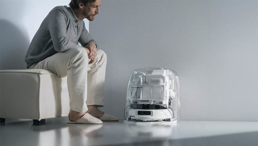 Osoba siedzi na kanapie, patrząc na przezroczysty robot sprzątający na podłodze. Robot ma nowoczesny design z widocznymi elementami wewnętrznymi.