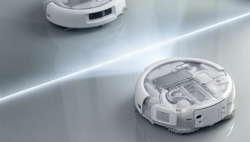 Na obrazie widoczne są dwa białe roboty sprzątające na gładkiej powierzchni. Roboty mają przezroczyste obudowy, ukazujące ich wewnętrzne mechanizmy.