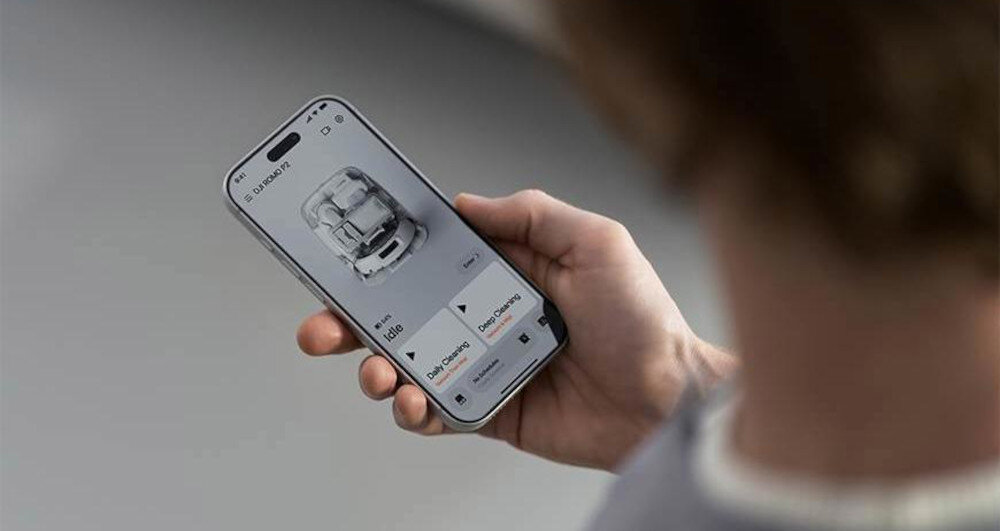 Osoba trzyma smartfon z aplikacją pokazującą obraz robota sprzątającego i opcje sterowania. Na ekranie widoczny jest tekst: \'DJI Romo P2 Clean More Combo\'.