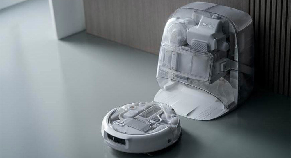 Na obrazie widoczny jest biały robot sprzątający oraz jego stacja dokująca na gładkiej powierzchni. Na stacji dokującej znajduje się napis \'ROMO\'.