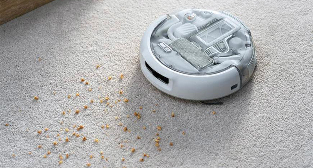 Robot sprzątający na jasnym dywanie zbiera rozsypane okruchy. Urządzenie ma okrągły kształt i przezroczystą górną pokrywę, przez którą widać jego mechanizmy.