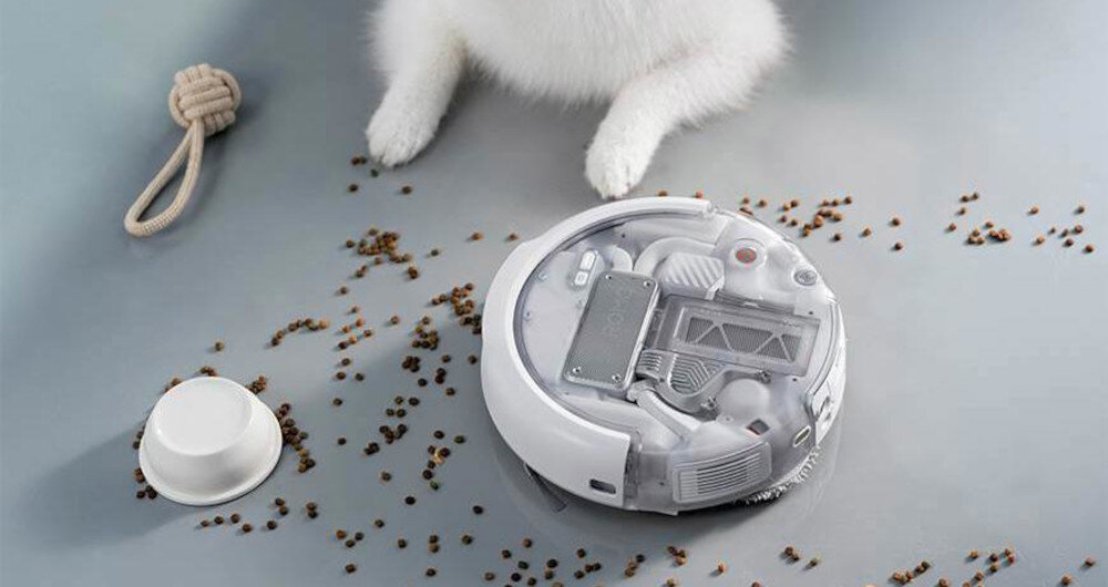 Na podłodze znajduje się okrągły robot sprzątający z przezroczystą pokrywą, obok którego leżą rozsypane drobne przedmioty. W tle widoczne są łapy białego zwierzęcia oraz zabawka w kształcie węzła.