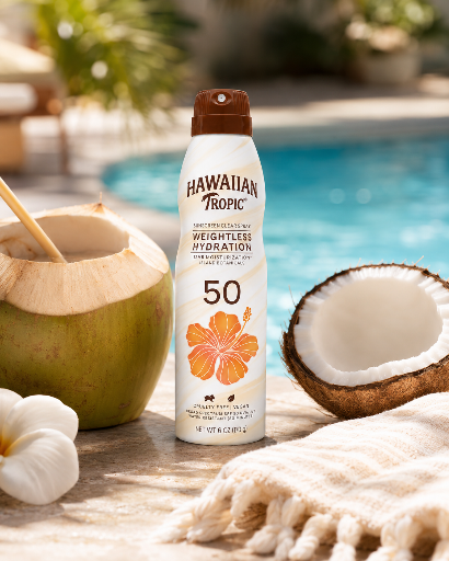 Na zdjęciu znajduje się spray do opalania Hawaiian Tropic SPF 50, obok którego leżą dwa kokosy i ręcznik, a w tle widać basen.