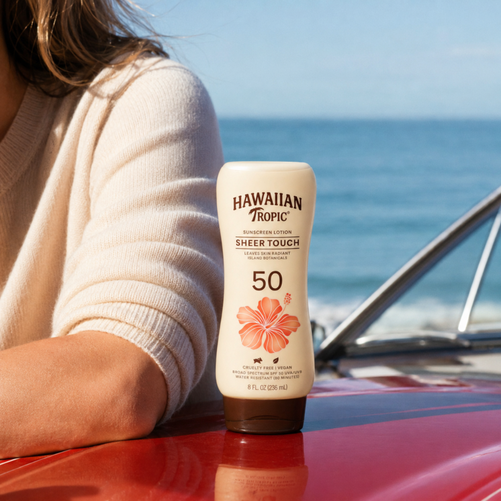 Na obrazie widoczna jest butelka kremu przeciwsłonecznego z napisem \'HAWAIIAN TROPIC SHEER TOUCH 50\'. W tle znajduje się morze i fragment samochodu.