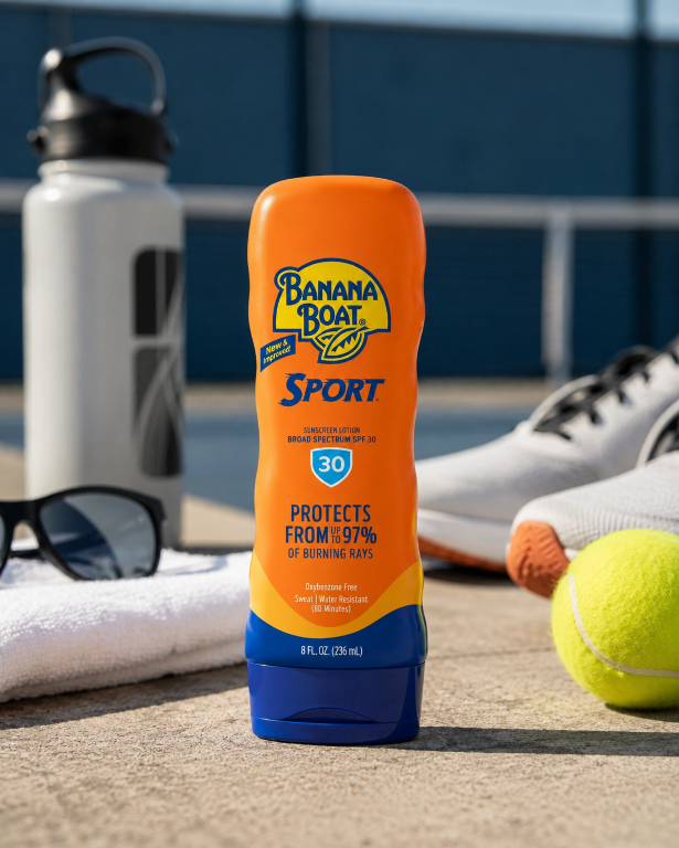 Na obrazie widoczna jest pomarańczowa butelka kremu przeciwsłonecznego Banana Boat Sport SPF 30, otoczona przez akcesoria sportowe, takie jak butelka na wodę, ręcznik, okulary przeciwsłoneczne, but sportowy i piłka tenisowa. Na butelce znajduje się tekst: \'PROTECTS FROM UP TO 97% OF BURNING RAYS\' oraz \'8 FL. OZ. (236 mL)\'.