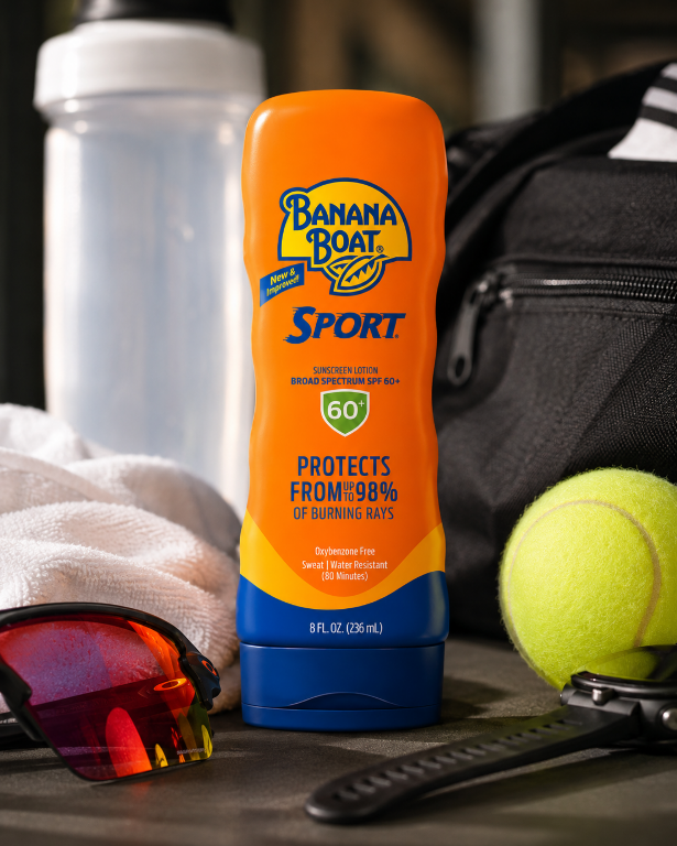 Na zdjęciu znajduje się pomarańczowa butelka balsamu do opalania z napisem \'Banana Boat Sport Sunscreen Lotion Broad Spectrum SPF 60+ 60+ Protects From 98% of Burning Rays Oxybenzone Free Sweat & Water Resistant (80 Minutes) 8 FL OZ (236 mL)\'. W tle widoczne są okulary przeciwsłoneczne, ręcznik, piłka tenisowa, butelka na wodę i torba sportowa.