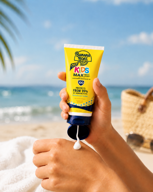 Osoba trzyma tubkę balsamu przeciwsłonecznego Banana Boat Kids Max Protect & Play SPF 100, z której wyciska się krem. W tle widać plażę i morze.