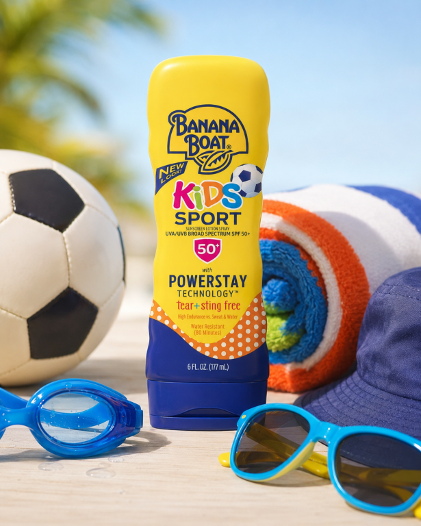 Na obrazie znajduje się żółta butelka balsamu z filtrem przeciwsłonecznym Banana Boat Kids Sport SPF 50+, otoczona przez piłkę, ręcznik, okulary przeciwsłoneczne i czapkę. Na butelce widnieje napis: \'Banana Boat Kids Sport Sunscreen Lotion Spray UVA/UVB Broad Spectrum SPF 50+ with Powerstay Technology tear + sting free\'.