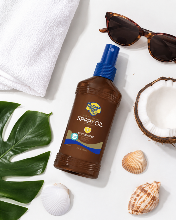 Na obrazie znajduje się brązowa butelka z niebieskim dozownikiem, oznaczona jako \'Banana Boat Deep Tanning Spray Oil 4 with Coconut Oil\'. Wokół butelki leżą okulary przeciwsłoneczne, ręcznik, połówka kokosa, muszle i zielony liść.