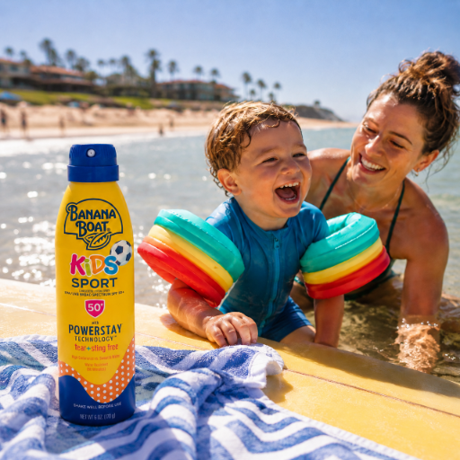Na plaży znajduje się dziecko w niebieskim stroju kąpielowym z kolorowymi rękawkami, obok dorosła osoba w wodzie. Na pierwszym planie widoczny jest spray do opalania Banana Boat Kids Sport SPF 50.