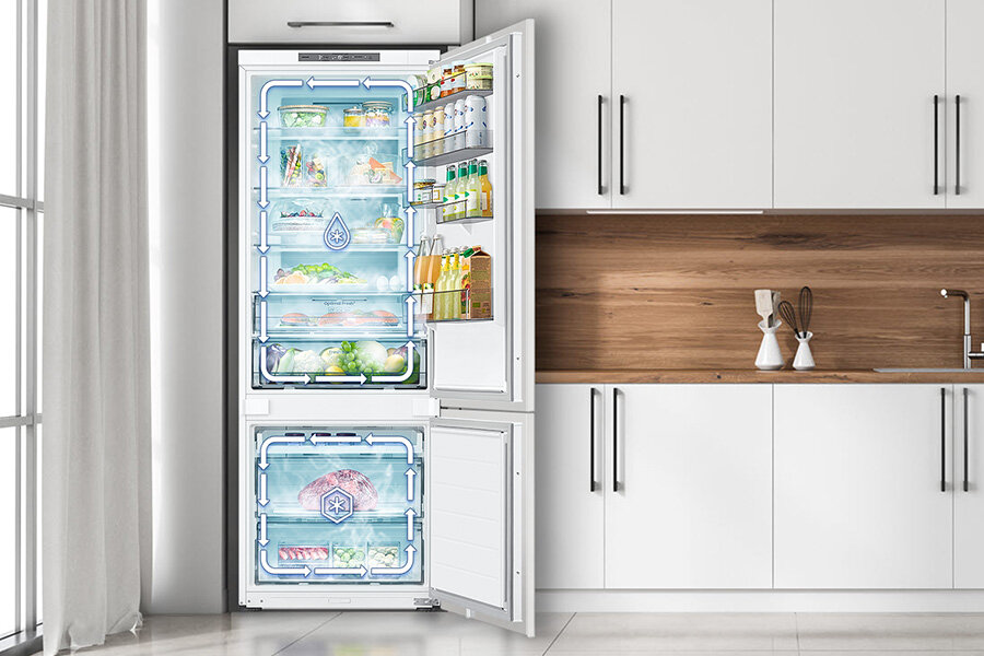 Otwarta lodówka z widocznymi półkami wypełnionymi różnymi produktami spożywczymi, takimi jak warzywa, napoje i mięso. Lodówka znajduje się w nowoczesnej kuchni z białymi szafkami i drewnianym blatem.