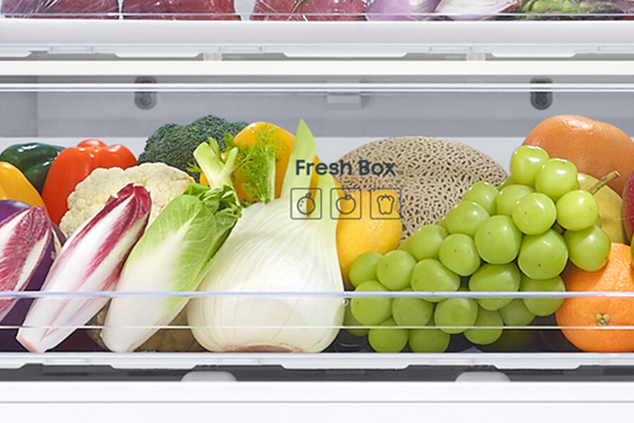 W lodówce znajduje się szuflada z różnymi warzywami i owocami, takimi jak winogrona, sałata, brokuły i papryka. Na szufladzie widoczny jest napis \'Fresh Box\' oraz ikony owoców i warzyw.