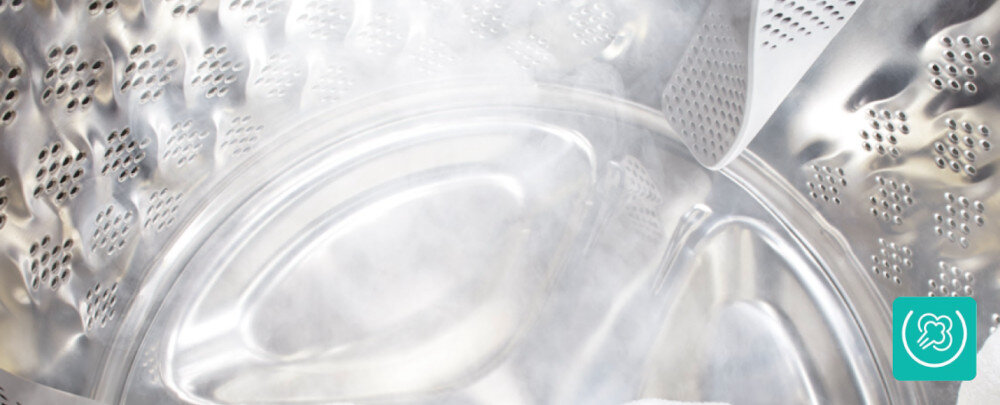 Wnętrze bębna pralki z widocznymi otworami i metaliczną powierzchnią. W prawym dolnym rogu znajduje się zielona ikona z symbolem prania.