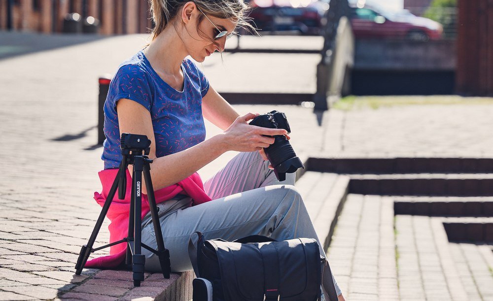 Osoba siedzi na schodach z aparatem fotograficznym, obok niej stoi czarny statyw. W tle widoczne są schody i zaparkowane samochody.