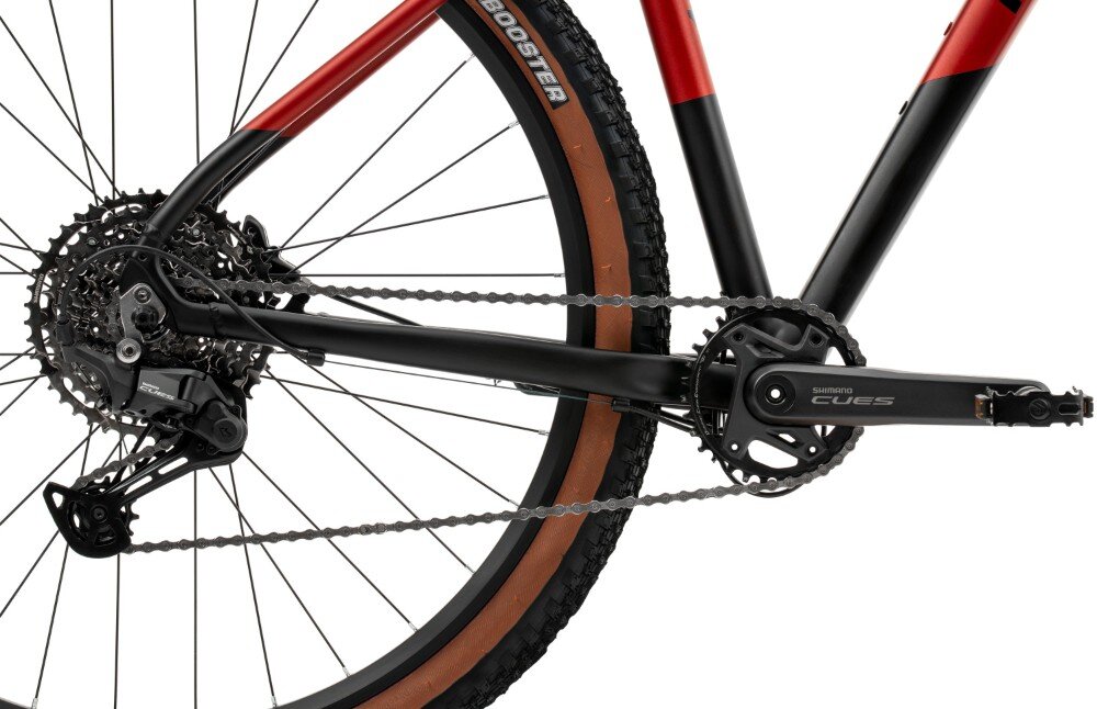 Zbliżenie na tylną część roweru górskiego z widocznym mechanizmem napędowym i kołem. Na oponie widoczny jest napis \'BOOSTER\', a na ramieniu mechanizmu napis \'SHIMANO CUES\'.