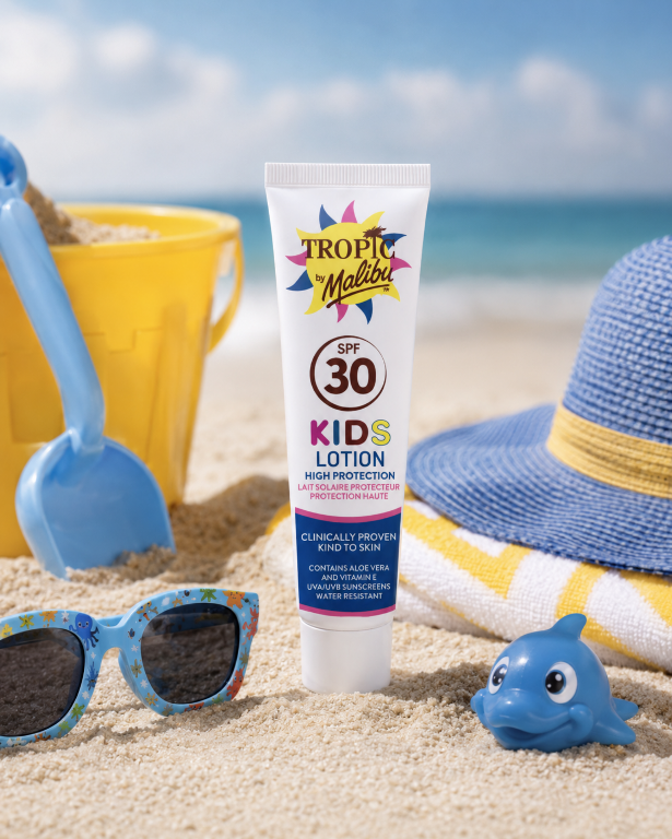 Na plaży znajduje się tubka balsamu \'Tropic by Malibu SPF 30 Kids Lotion High Protection\'. W tle widoczne są akcesoria plażowe, takie jak wiaderko, łopatka, kapelusz, okulary przeciwsłoneczne i zabawka w kształcie delfina.