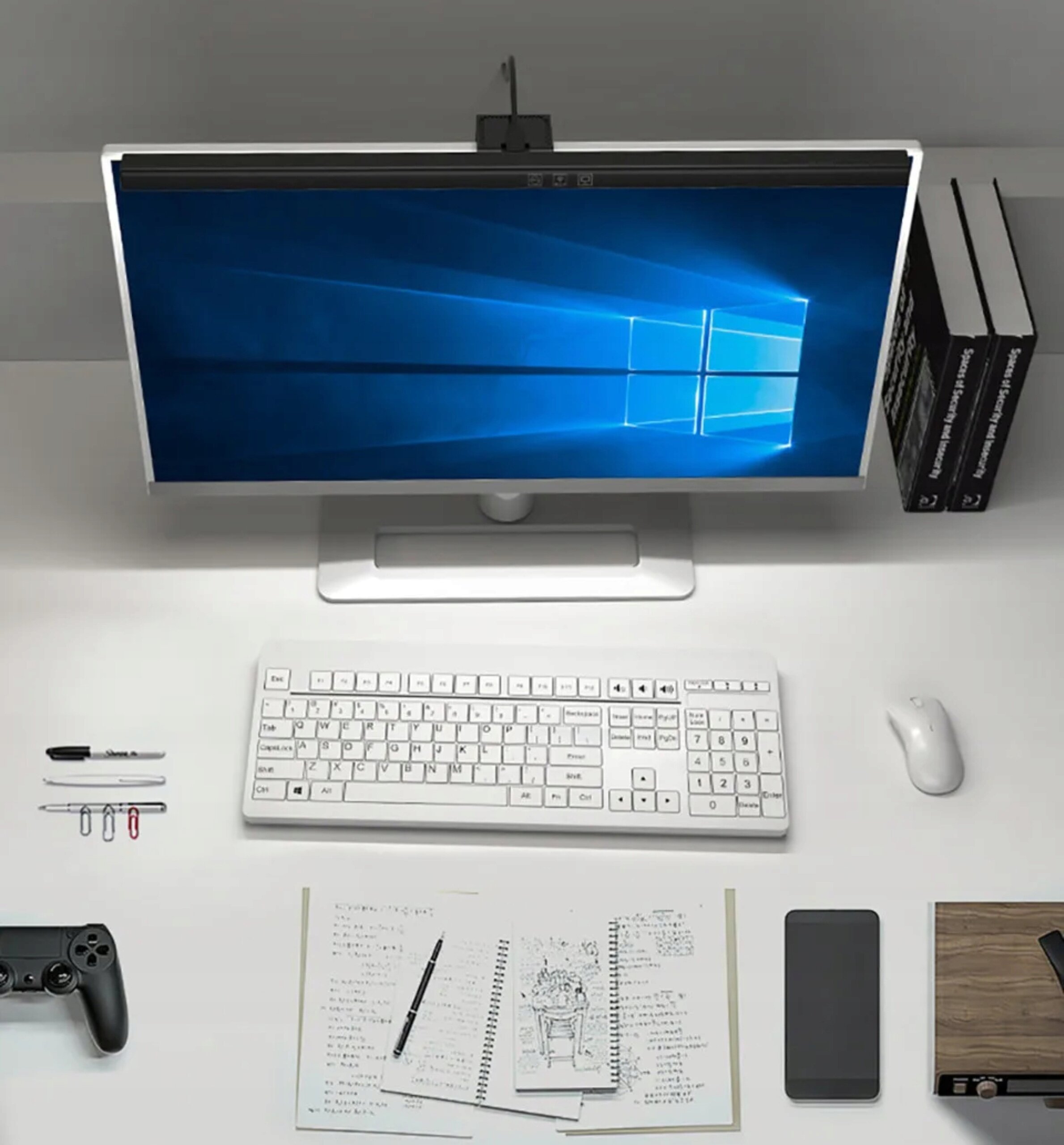 Na biurku znajduje się monitor z włączonym systemem Windows, przed nim klawiatura i mysz. Obok leży kontroler do gier, długopisy, notatki oraz smartfon.