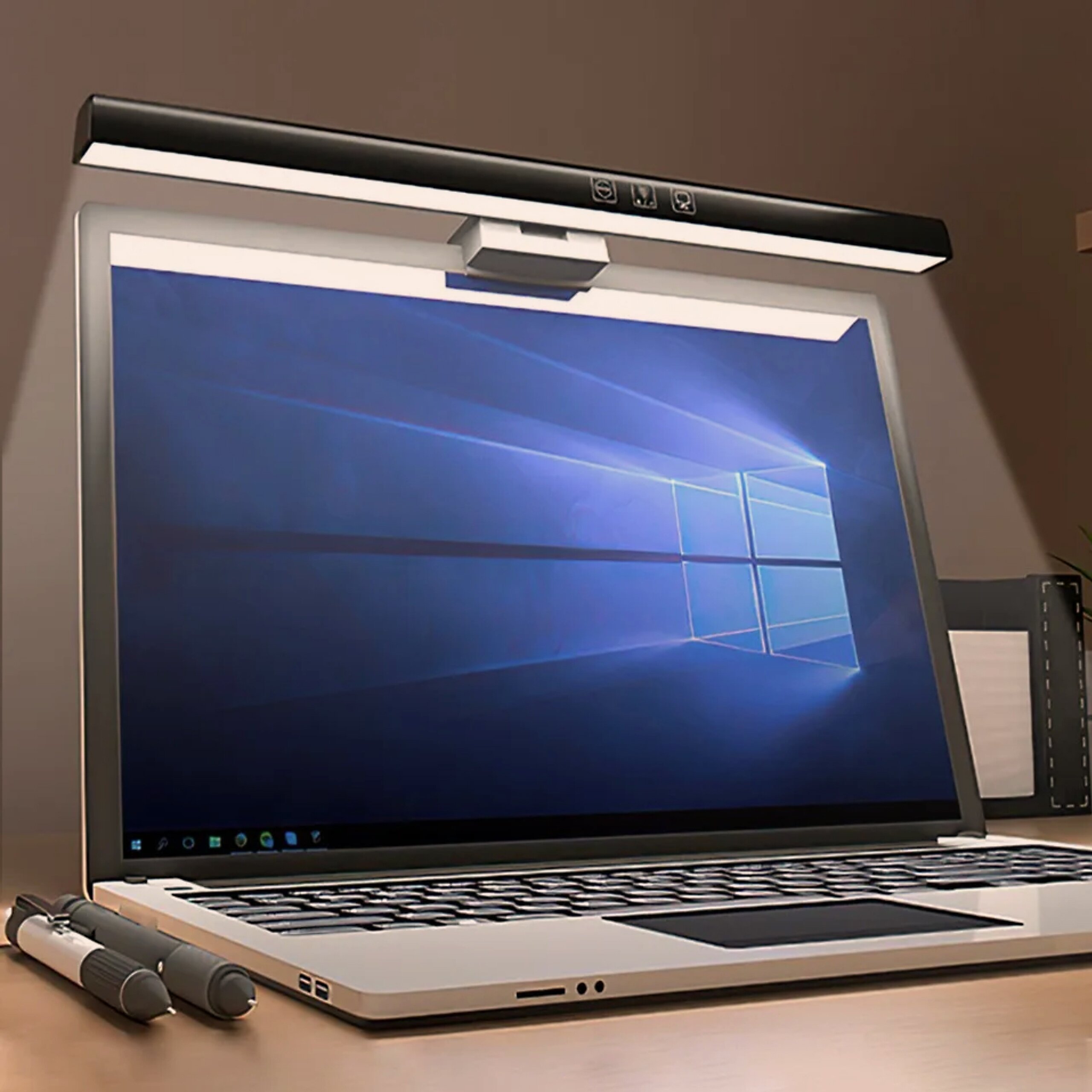 Na obrazie widać laptop z włączonym ekranem, nad którym zamontowana jest lampka LED. Na biurku obok laptopa leżą dwa długopisy i notes.
