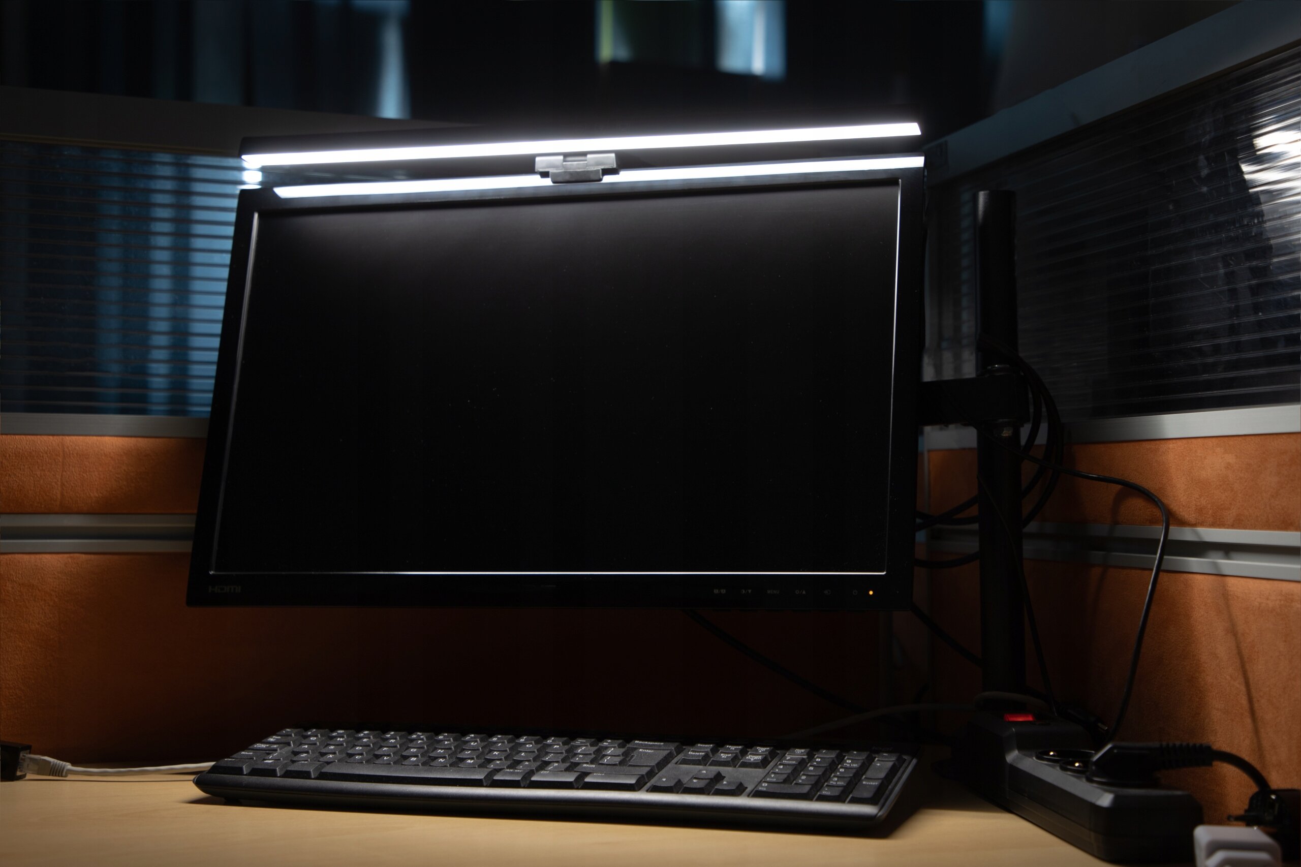 Na biurku znajduje się monitor z zamontowaną na górze lampką LED. Przed monitorem leży klawiatura, a obok widoczny jest przedłużacz z kilkoma podłączonymi kablami.