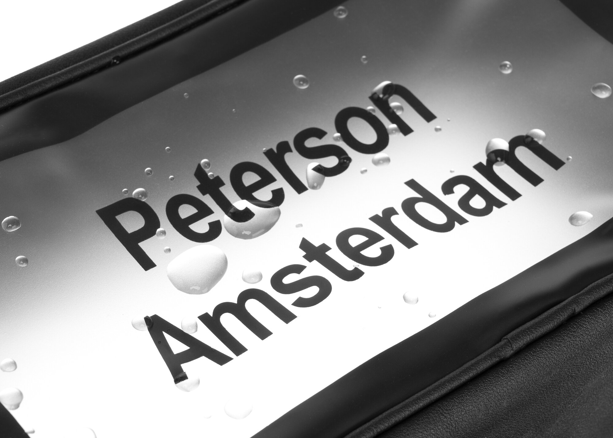 Na obrazie widoczny jest fragment przezroczystej powierzchni z czarnym napisem \'Peterson Amsterdam\', na której znajdują się krople wody.