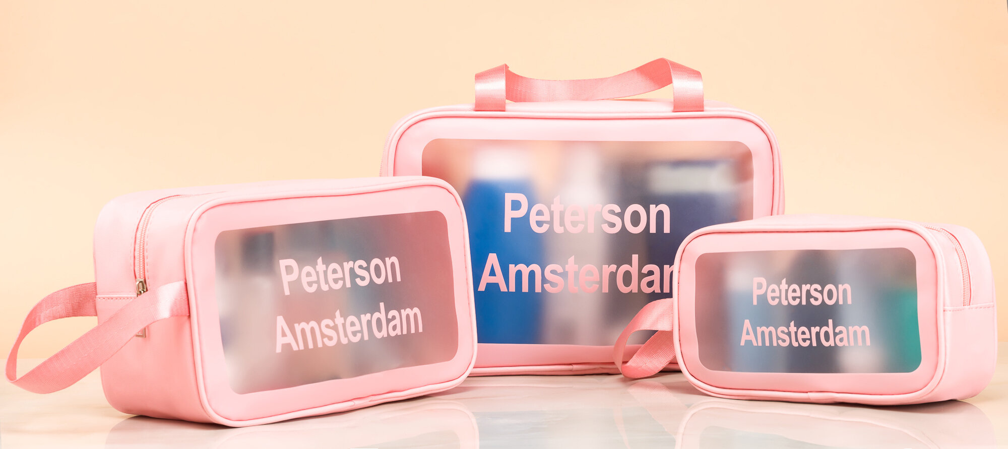 Trzy różowe kosmetyczki z przezroczystymi panelami, na których widnieje napis \'Peterson Amsterdam\'. Kosmetyczki mają różne rozmiary i różowe uchwyty.