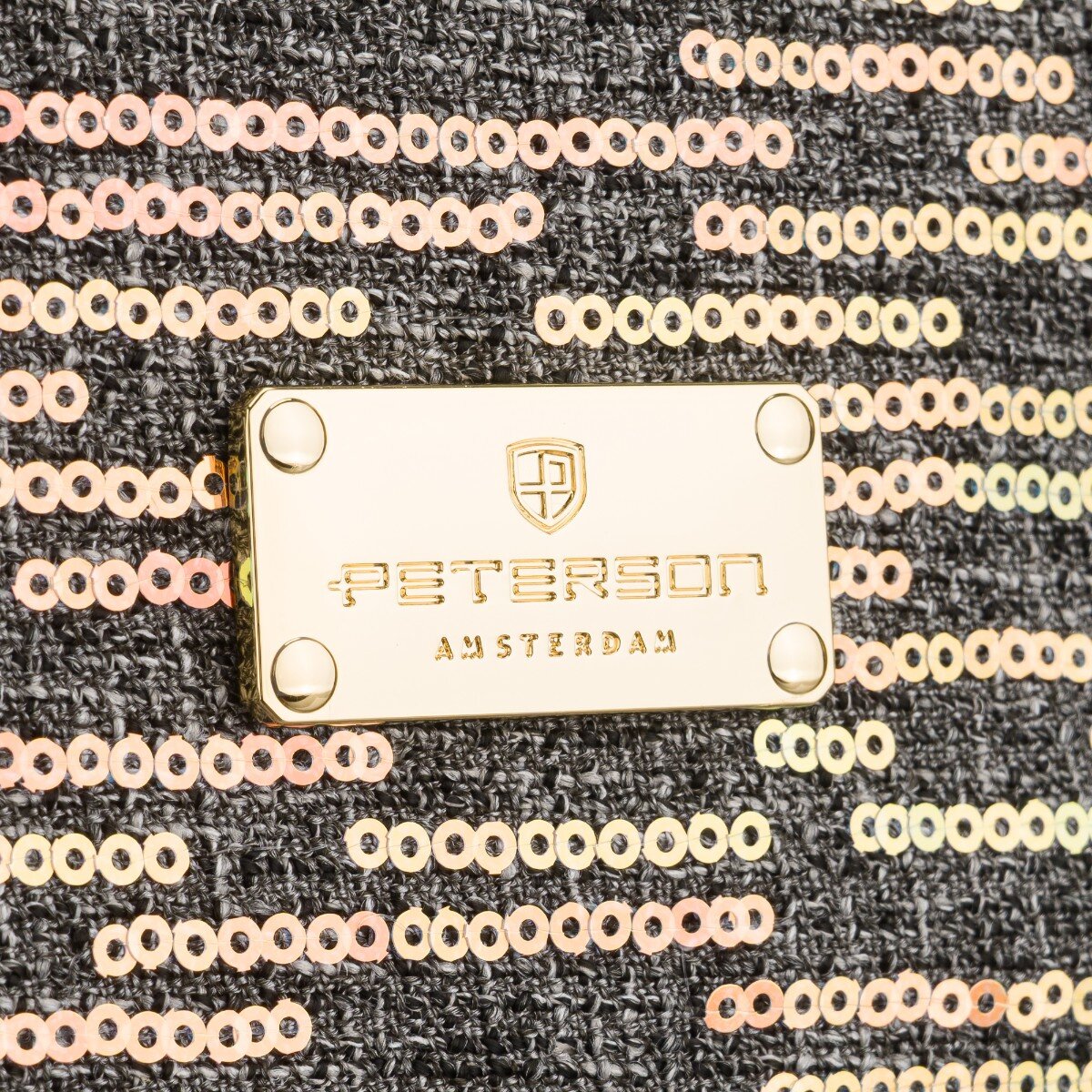 Zbliżenie na czarny materiał pokryty cekinami z metalową plakietką. Na plakietce widnieje napis: \'PETERSON AMSTERDAM\'.
