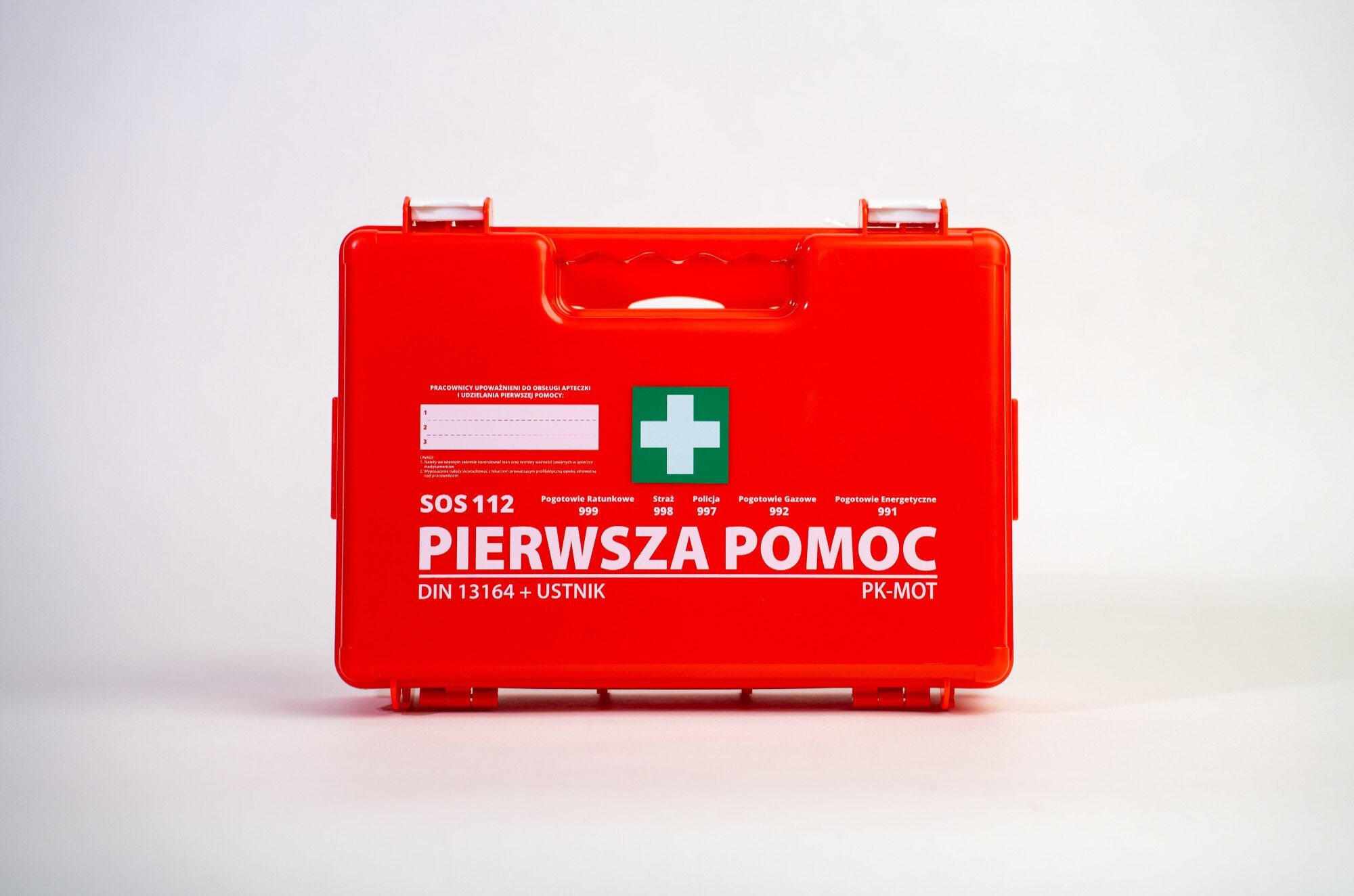 Czerwona apteczka z białym krzyżem na zielonym tle oraz napisem \'PIERWSZA POMOC\'. Widoczne są również numery alarmowe i oznaczenie \'DIN 13164 + USTNIK\'.