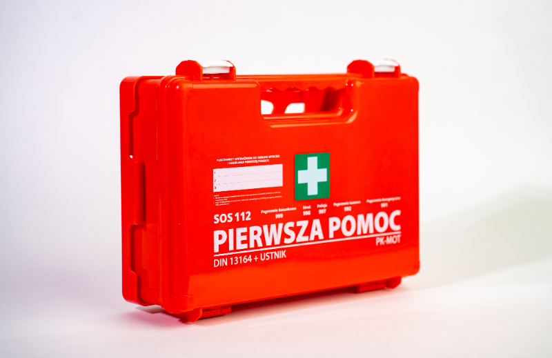 Czerwona apteczka z białym krzyżem na zielonym tle oraz napisem \'PIERWSZA POMOC\' i \'DIN 13164 + USTNIK\'.