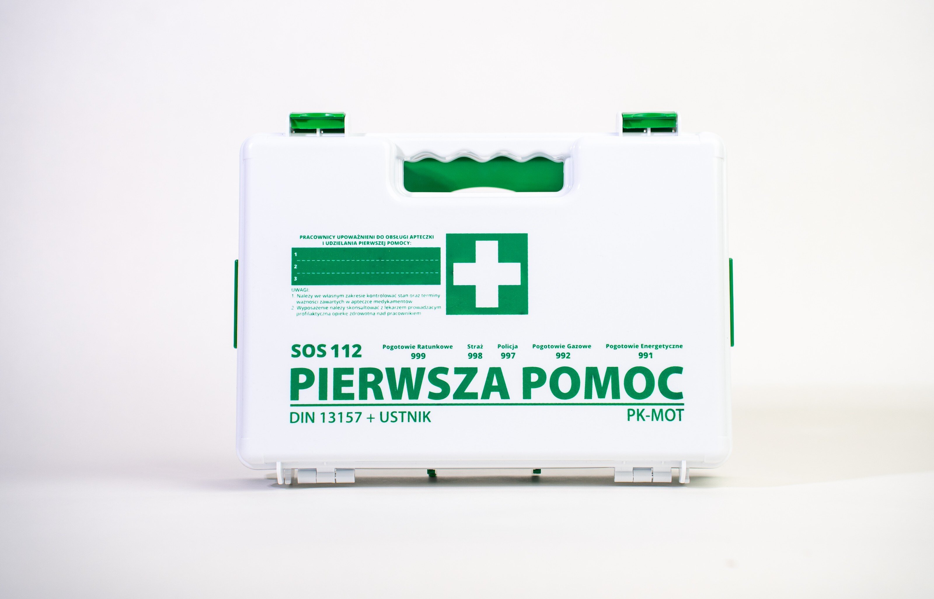 Biała apteczka z zielonym uchwytem i napisem \'PIERWSZA POMOC\' oraz symbolem krzyża. Widoczne są numery alarmowe i oznaczenie DIN 13157 + USTNIK.